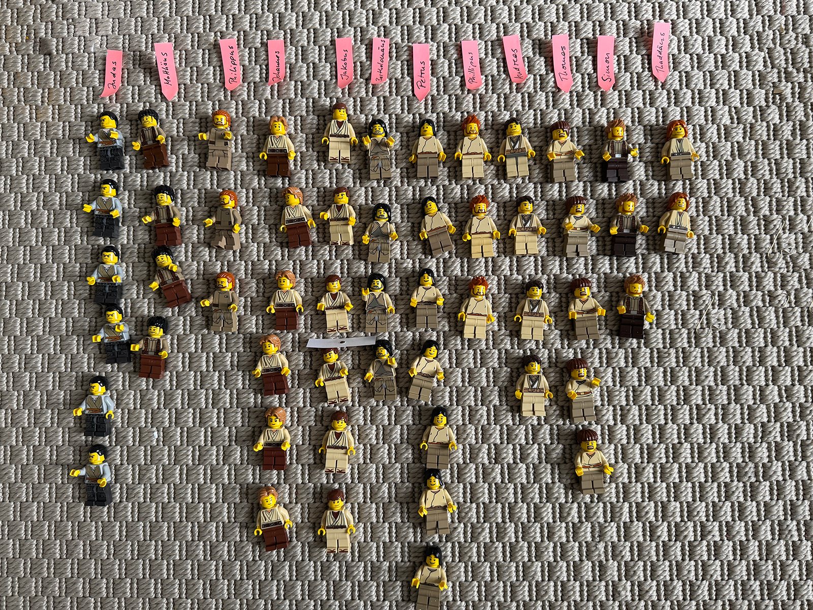 Dutzende Apostel-Minifiguren in Papiergewändern liegen auf einem Teppich, nach Namen sortiert, darüber kleine rosa Namenszettel