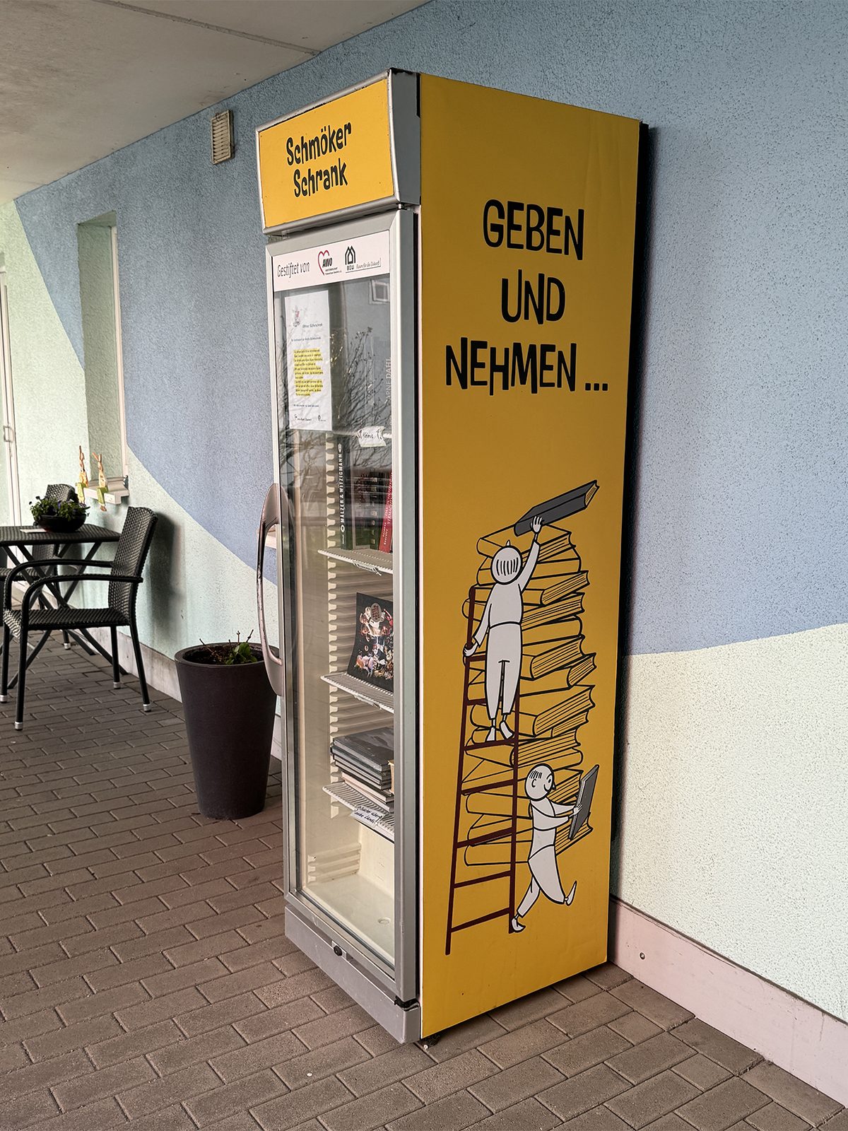Gelber „Schmöker Schrank“ vor hellblauer Wand: Auf der Seite steht „Geben und nehmen …“, darunter zwei gezeichnete Figuren an einer Leiter vor einem Bücherstapel