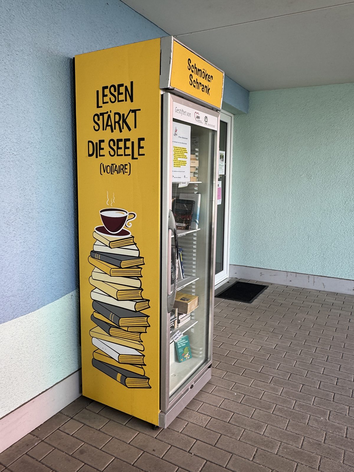Die andere Seite des gelben Schranks: „Lesen stärkt die Seele (Voltaire)“, darunter ein Bücherstapel mit einer Kaffeetasse