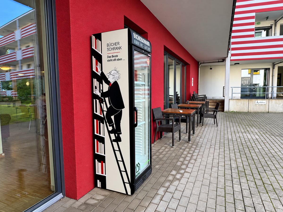Seitenansicht des Bücherschranks vor der roten Hauswand: Ein gezeichneter Mann steigt eine Leiter am Bücherregal hinauf, darüber „Das Beste steht oft oben …“