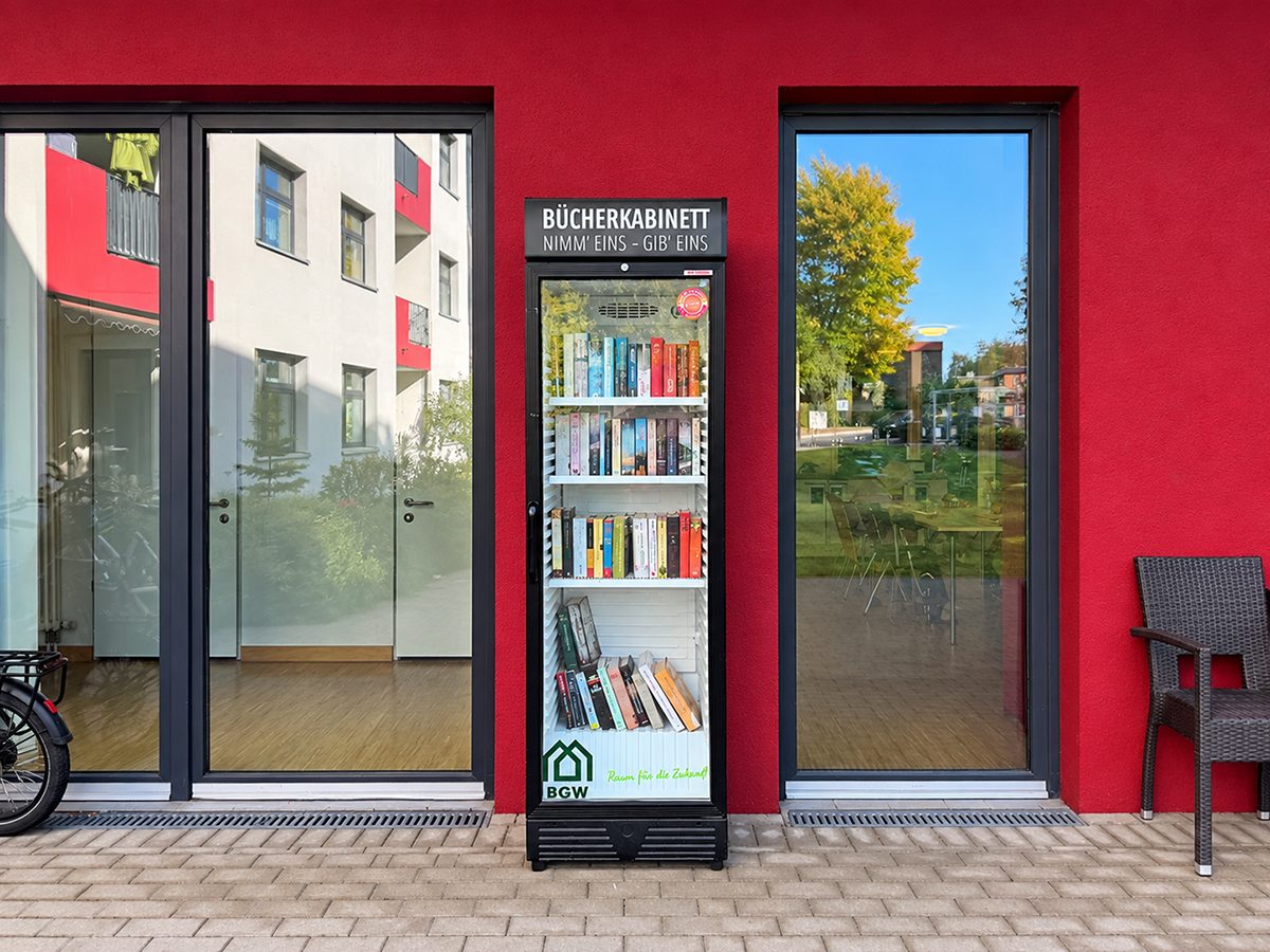 Frontansicht: Der Bücherschrank „Bücherkabinett“ zwischen zwei Glastüren vor der roten Hauswand, gefüllt mit bunten Büchern