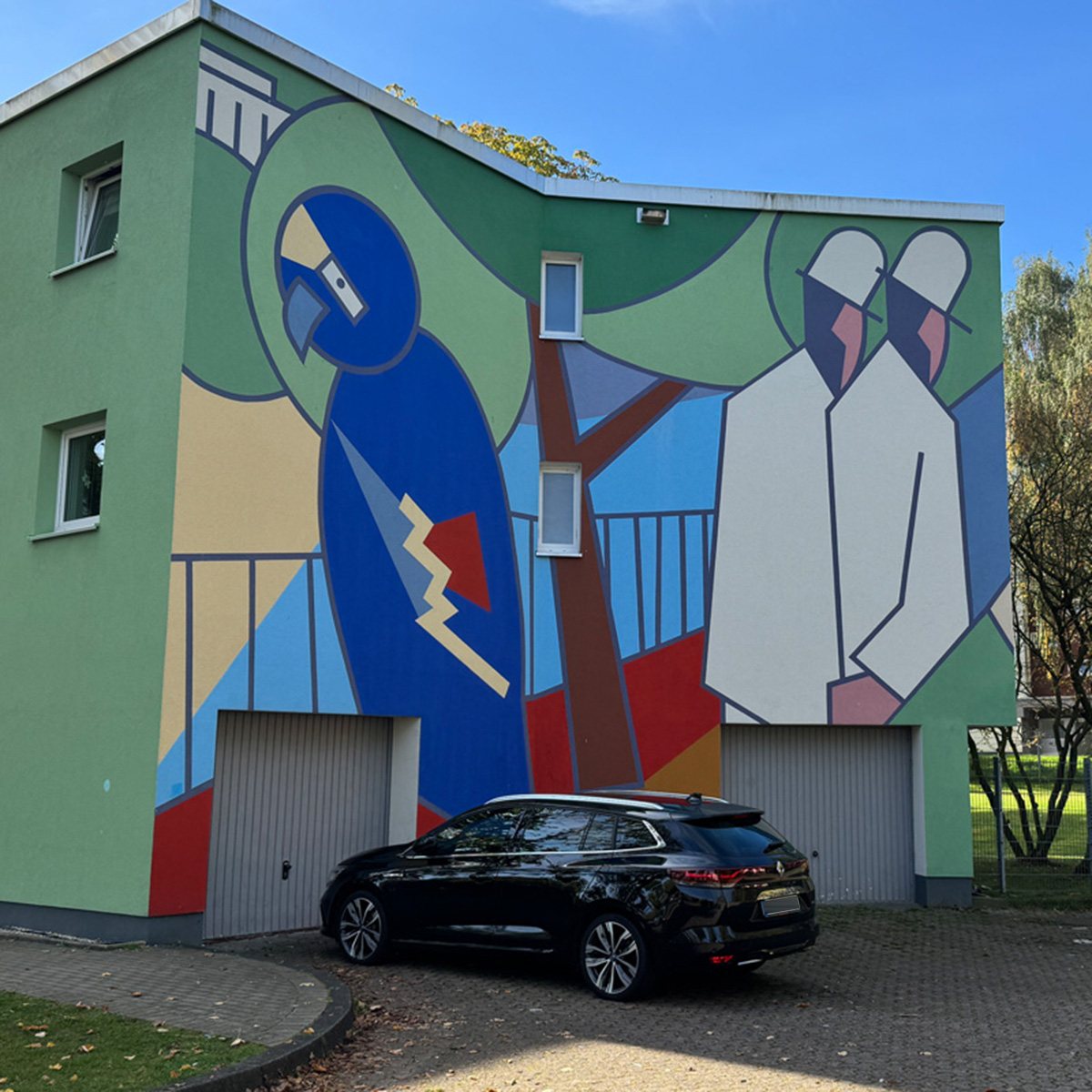 Fassadenbild im Rabenhof: Figuren in Blau, Weiß und Rot auf grünem Grund an der Stirnseite eines Hauses mit Garagen