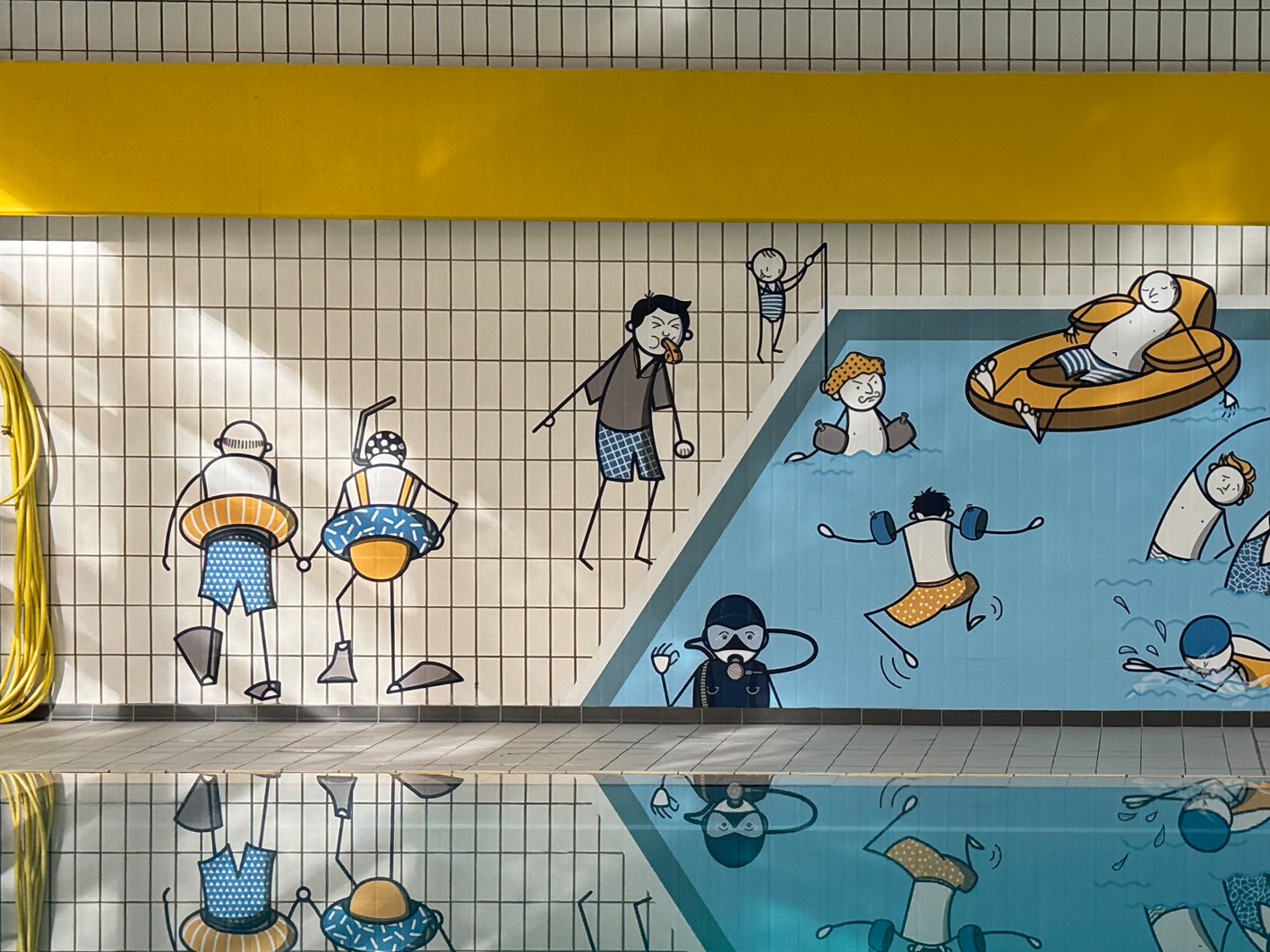 Ausschnitt einer Wandgestaltung im Hallenbad: gezeichnete Badegäste mit Schwimmreifen, Bademeister mit Trillerpfeife und Taucher auf weißen und hellblauen Fliesen, gespiegelt im Wasser