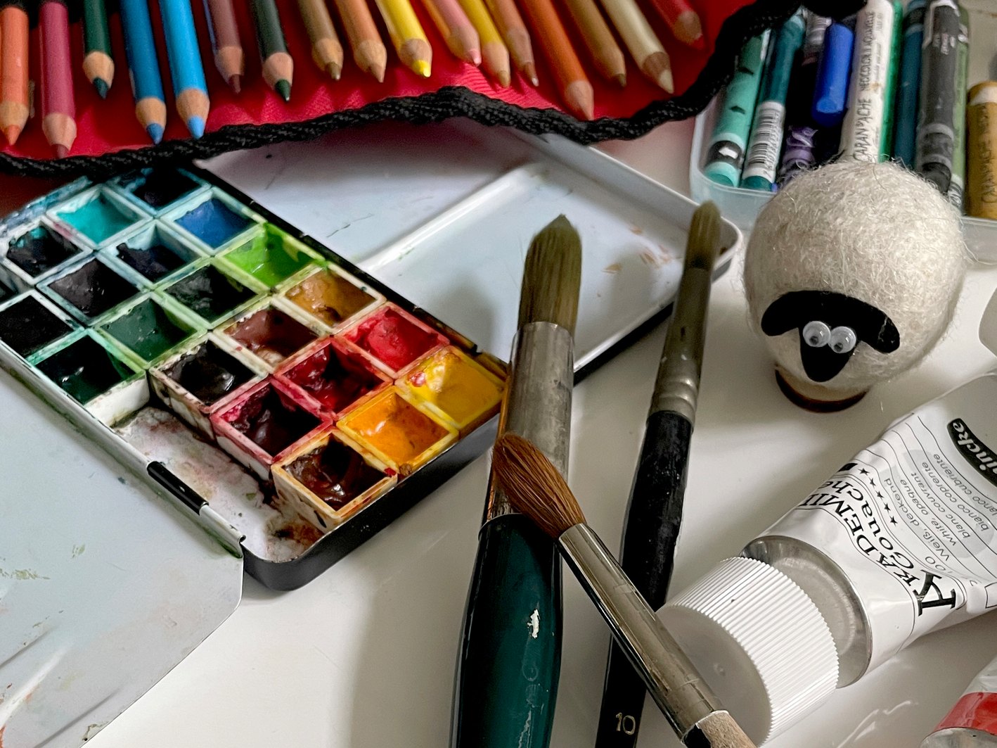 Arbeitsmaterial auf dem Tisch: Aquarellkasten, Pinsel, Buntstifte, Ölkreiden, eine Tube Gouache und ein kleines Filzschaf