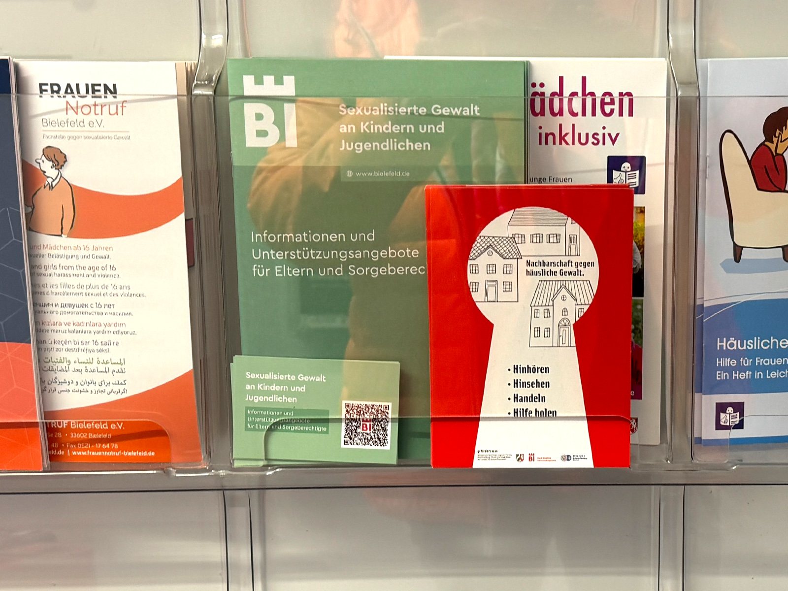 Foto: Die rote Postkarte „Nachbarschaft gegen häusliche Gewalt.“ steckt in einem Prospektständer zwischen anderen Informationsflyern