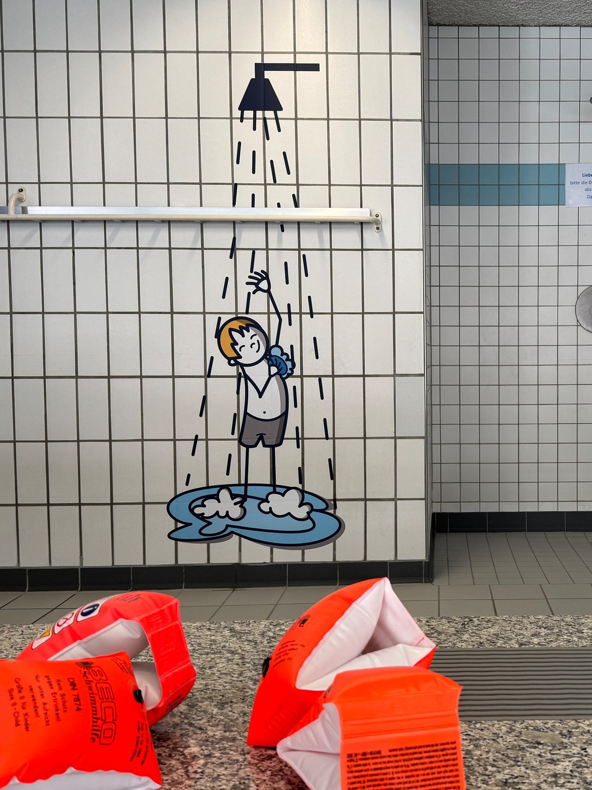 Gezeichneter Junge unter der Dusche an der Fliesenwand, davor echte Schwimmflügel