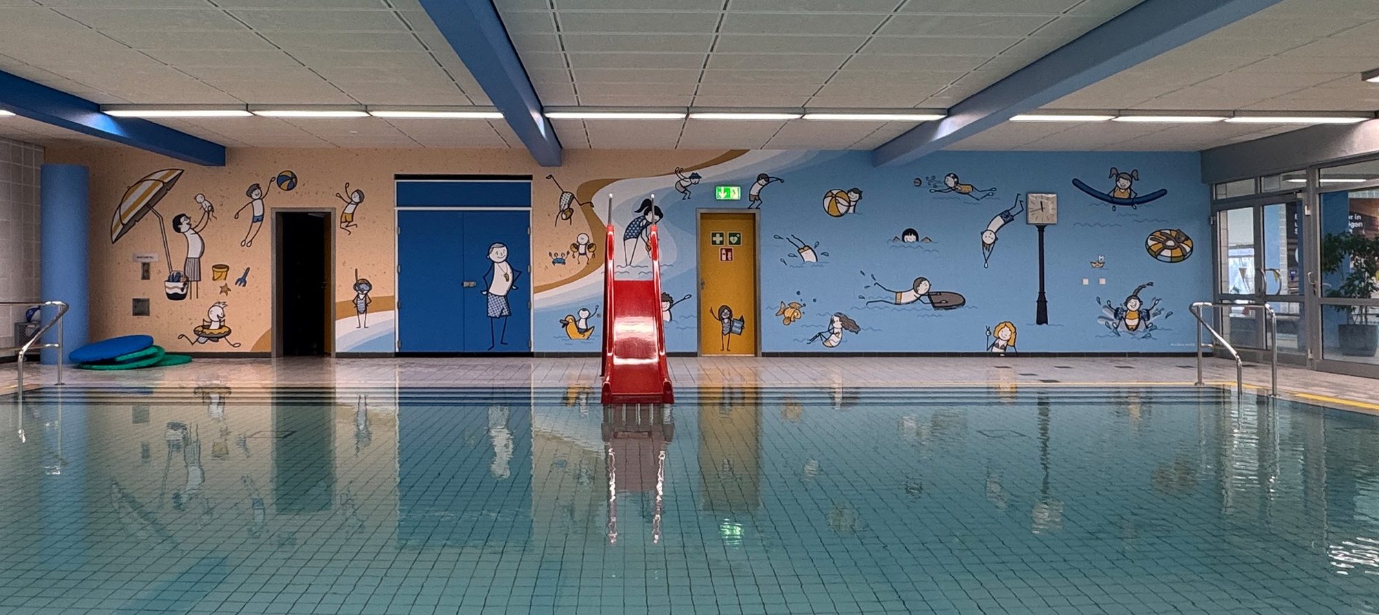 Gesamtansicht des Lehrbeckens mit roter Rutsche und der umlaufend illustrierten Wand, gespiegelt im Wasser