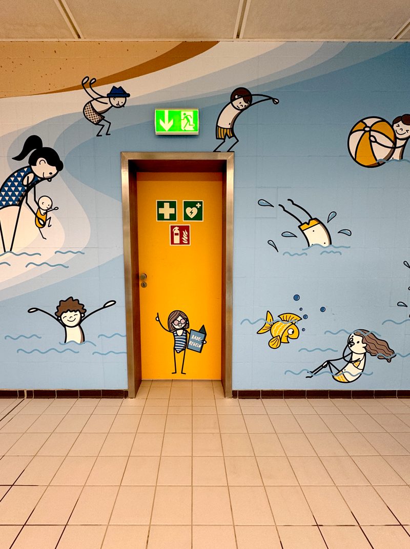 Gelbe Tür im Lehrbecken, umgeben von gezeichneten Badegästen; auf der Tür eine kleine Figur mit Buch