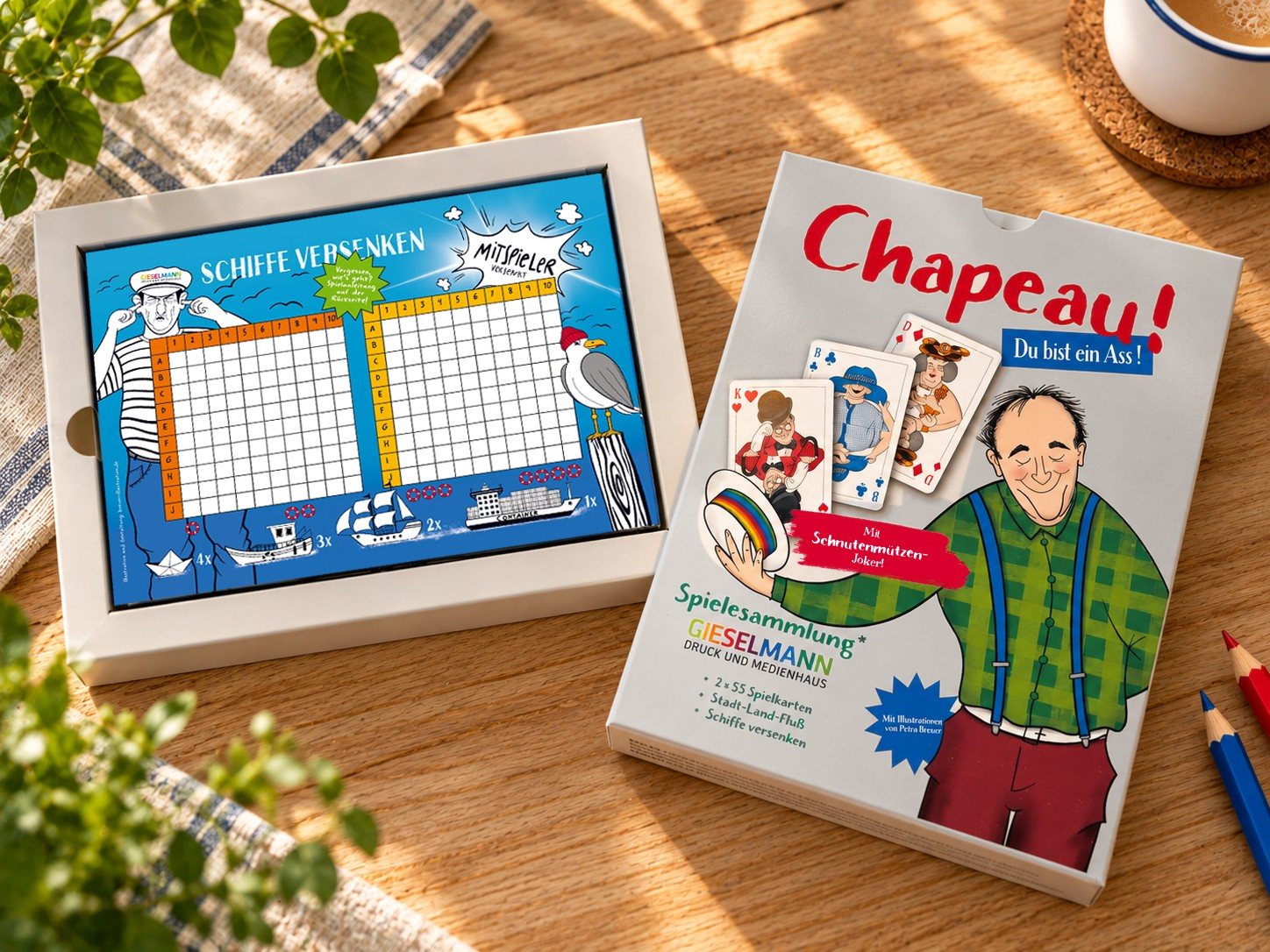 Die Spielesammlung auf einem Holztisch: links die geöffnete Schachtel mit dem Block „Schiffe versenken“, rechts der Schuber mit dem Titel „Chapeau!“