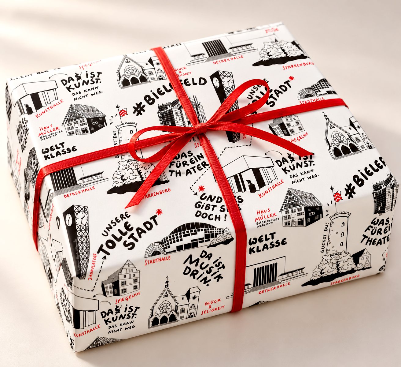 Ein Geschenk, eingepackt in schwarz-weißes Bielefeld-Papier mit roter Schleife; auf dem Papier Wahrzeichen und Sprüche wie „Unsere tolle Stadt“ und „#Bielefeld“